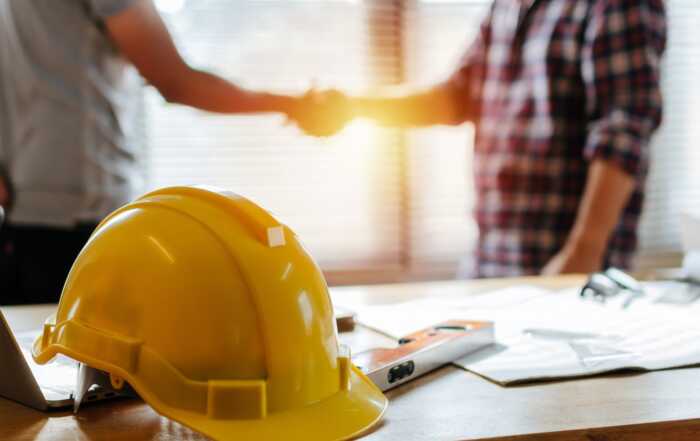 Yellow safety helmet on workplace desk with construction worker team hands shaking greeting start up plan new project contract in office center at construction site, partnership and contractor concept stock photo