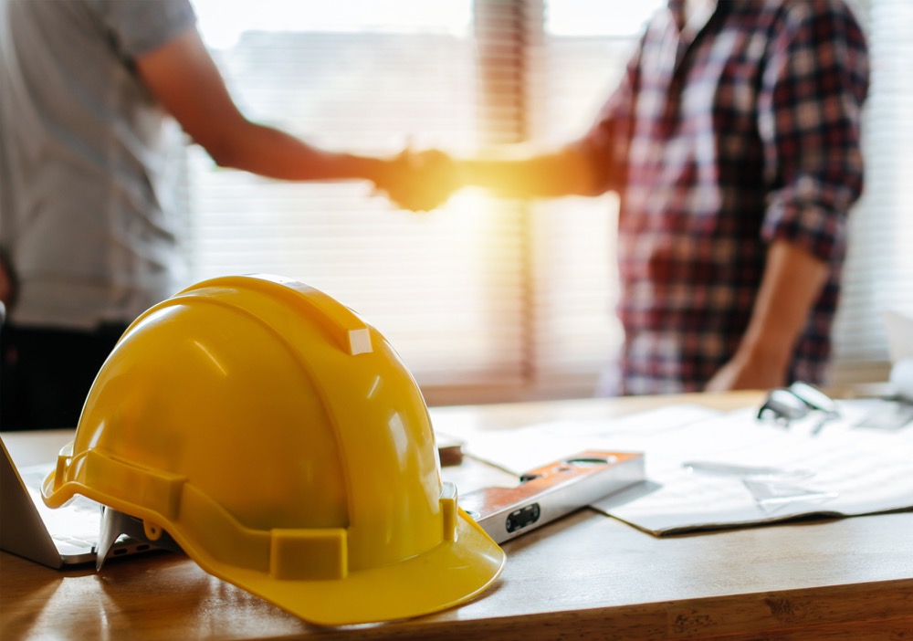 Yellow safety helmet on workplace desk with construction worker team hands shaking greeting start up plan new project contract in office center at construction site, partnership and contractor concept stock photo